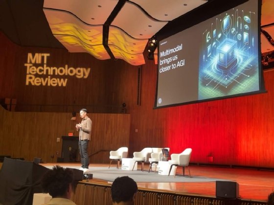 A man stands onstage with 'MIT Technology Review' projected on the wall. Behind him, a presentation slide reads 'Multimodal brings us closer to AGI.'